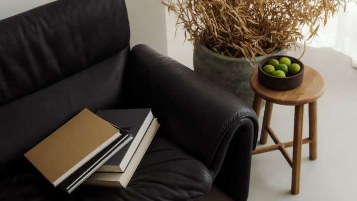 High-angle shot of a minimalist, modern interior. A stack of notebooks with black and tan covers rests on the arm of a black leather sofa. Next to the sofa is a light-wood stool holding a small wooden bowl filled with bright green limes. Behind the stool is a large, textured planter containing dry, straw-like decorative foliage. The room is brightly lit with a white floor.