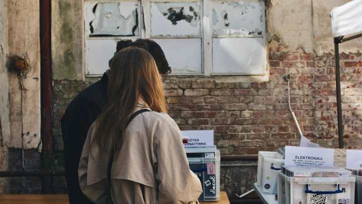A man and a woman stand with their backs to the camera, looking down at items on a table at an outdoor market or fair. The woman wears a tan trench coat. They are standing against a background of an old brick wall with a dilapidated, white-framed window above. The table in the foreground holds several white plastic bins.