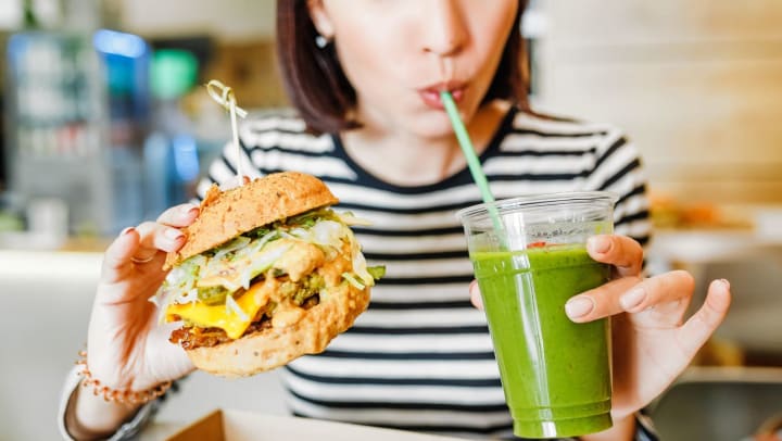 a woman sips green juice and holds a vegan burger with toppings on it at a vegan fast food place | vegan restaurants in Charlotte