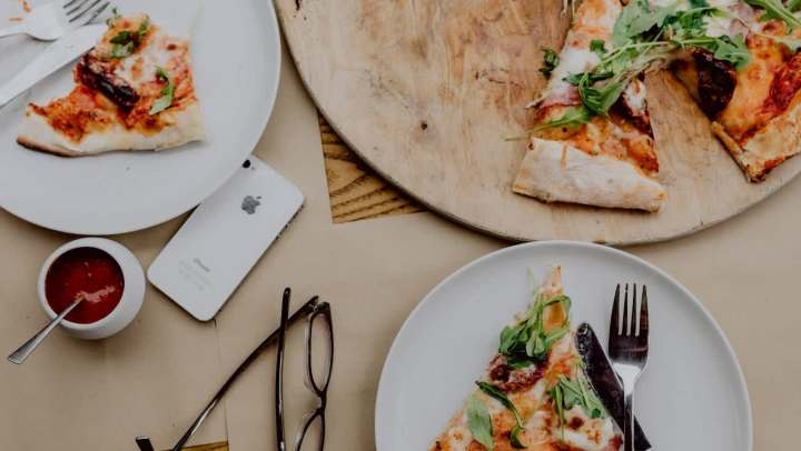 Flat-lay shot of a table set for a casual meal, featuring several slices of thin-crust pizza topped with arugula and prosciutto. The slices are on two white plates and a round wooden serving board. Also visible on the brown table surface are an iPhone, dark-framed eyeglasses, and small bowls of red and white dipping sauces.