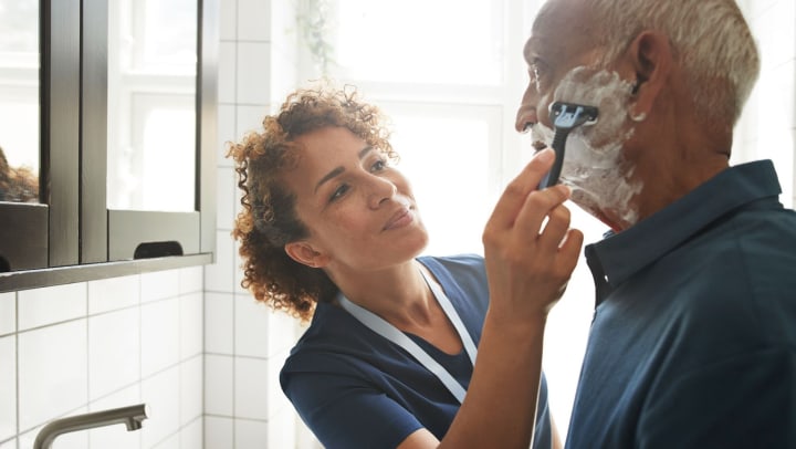 Healthcare worker shaving older man in a bathroom