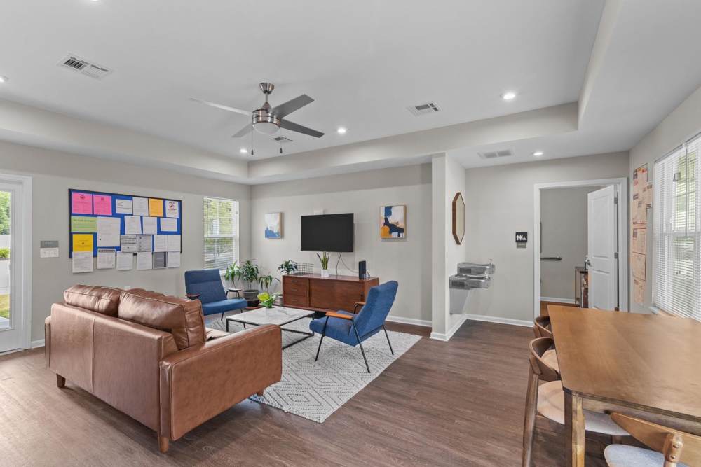 Modern community lounge with leather sofa, blue chairs, and wood-style flooring, bright natural light streaming in at Old Firetower Place in Greenville, North Carolina