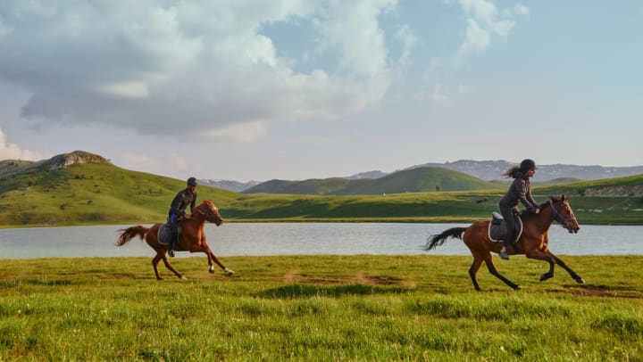 Two people riding horses along a lake