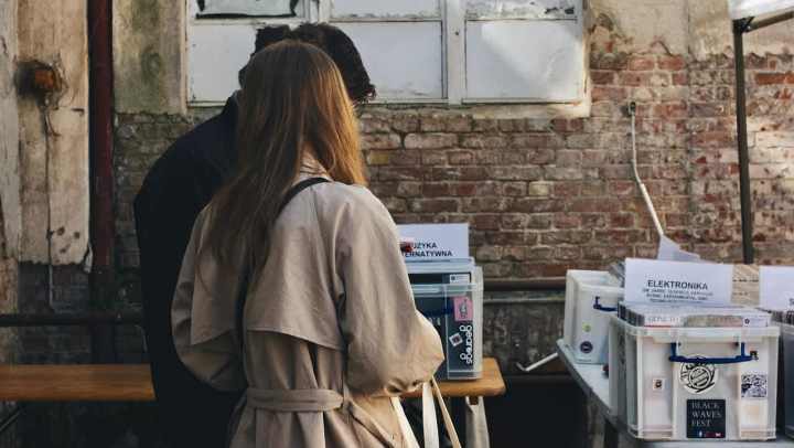 A man and a woman stand with their backs to the camera, looking down at items on a table at an outdoor market or fair. The woman wears a tan trench coat. They are standing against a background of an old brick wall with a dilapidated, white-framed window above. The table in the foreground holds several white plastic bins.