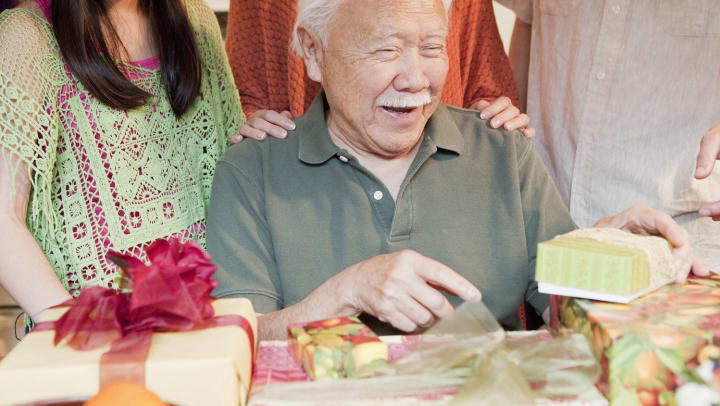 A smiling senior man is excited while opening presents from his family who are standing behind him.