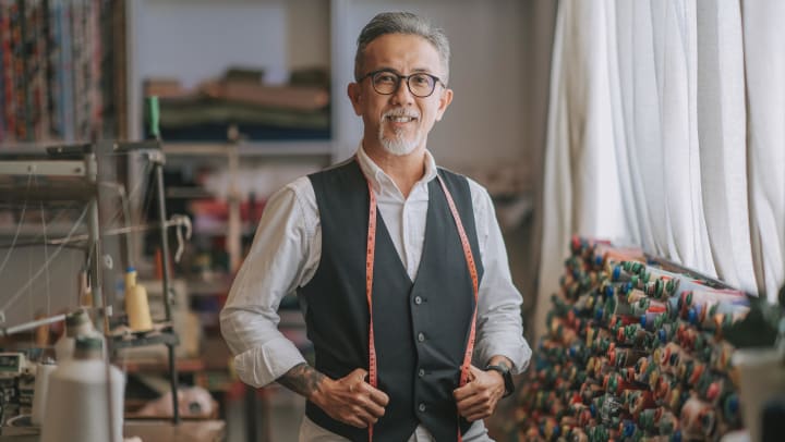 a sophisticated, gray-haired tailor wearing black-framed glasses smiles at the camera | tailors around gilbert