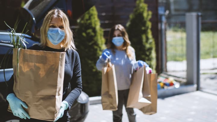 Two women wearing masks carrying groceries from a car, near The Slate