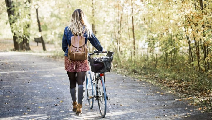 A woman wearing a denim jacket and a brown backpack walks her bike along a path in a wooded park setting | Charlotte parks