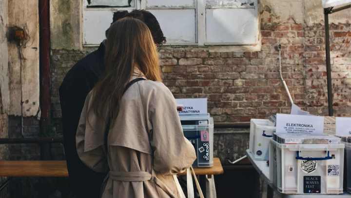 A man and a woman stand with their backs to the camera, looking down at items on a table at an outdoor market or fair. The woman wears a tan trench coat. They are standing against a background of an old brick wall with a dilapidated, white-framed window above. The table in the foreground holds several white plastic bins.