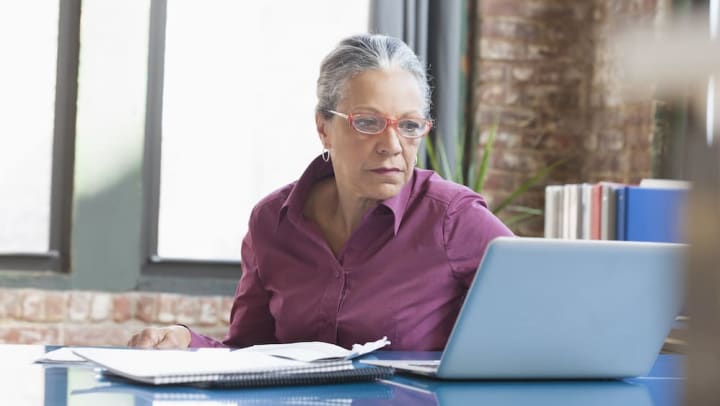 Older woman sitting at a desk with notebook and papers, looking at a laptop