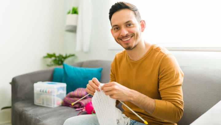 A man knitting a wool scarf and smiling at the camera.