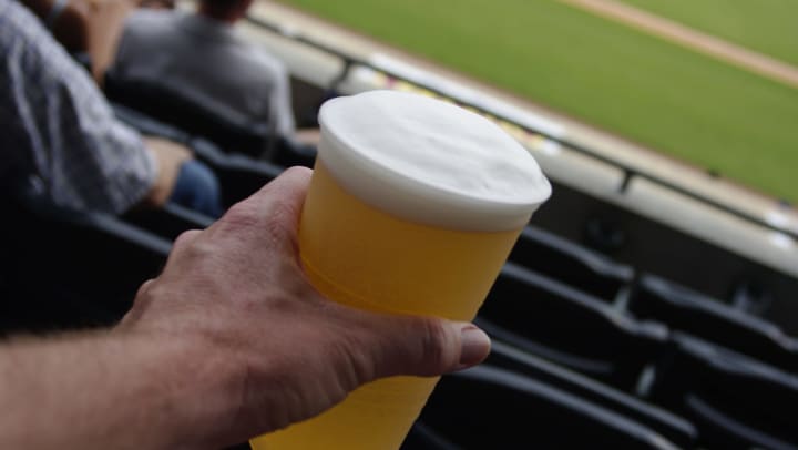 Closeup of man holding plastic cup of beer with baseball stadium blurred in background