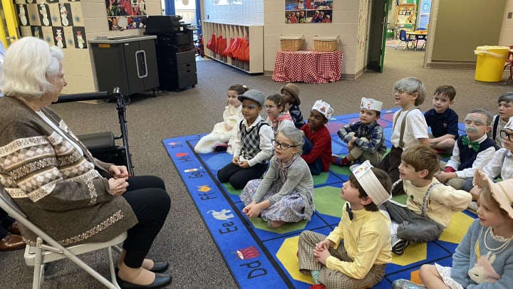 Peggy Holly is seated left speaking to a group of kindergarteners seated on a blue mat