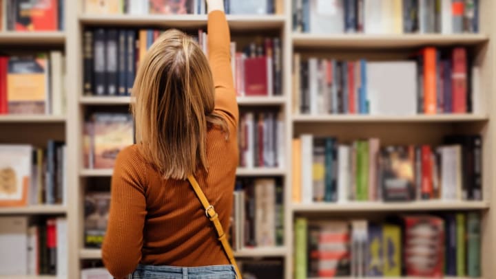 A shopper in an orange shirt reaches up to pull a paperback title from a top shelf