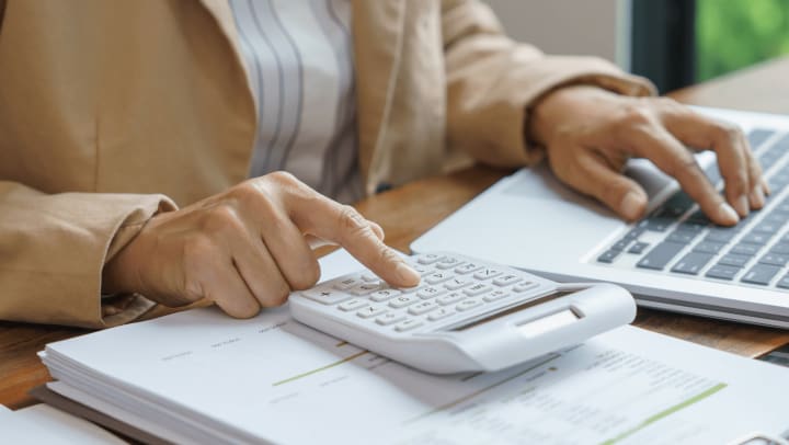 Person using a calculator and laptop to review financial documents, representing budgeting, financial planning, or economic analysis.