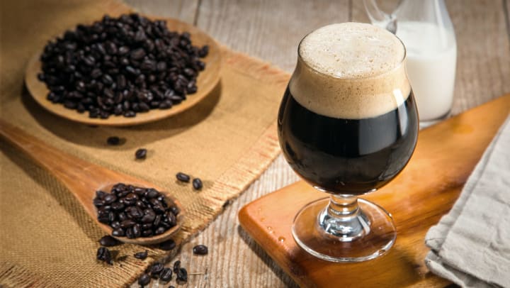 A snifter glass full of dark beer sitting on a wooden cutting board with a bowl of coffee beans and a pitcher of milk sitting near it. 