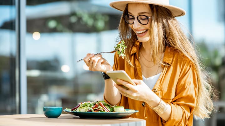 A smiling woman sitting at an outdoor table eats a salad as she looks at her phone.