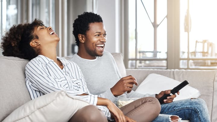 Young man and young woman sitting on a couch in a sunlit interior and laughing while watching television