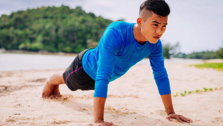 Young man in black shorts and blue long-sleeve shirt holding a plank pose on a sandy beach