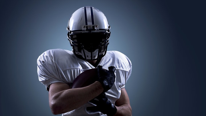 A football player in a white helmet and jersey, face covered in shadow, holding a football. 