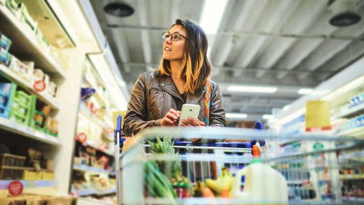 Woman with cart in grocery aisle consulting shopping list on her cell phone
