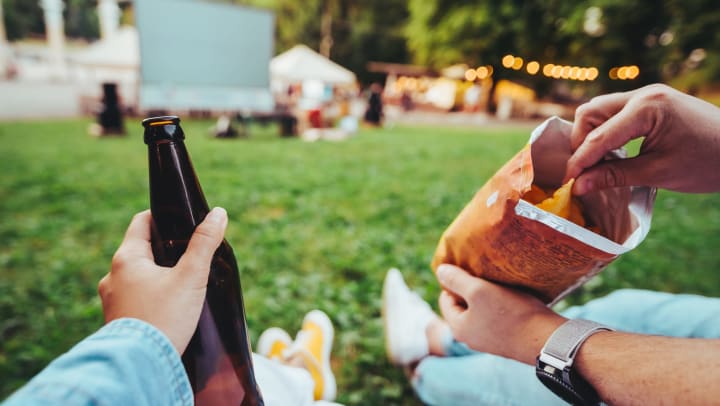 One person’s hand holding a glass bottle and another holding a bag of chips in an open-air cinema. 