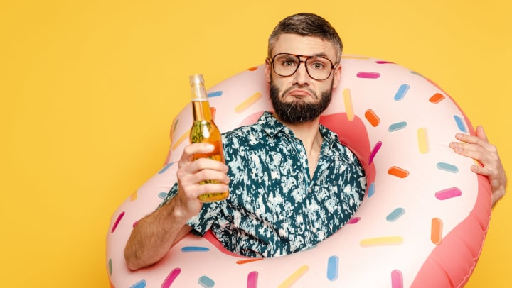 Man holding a beer, with a large pink inflatable donut over his shoulder.