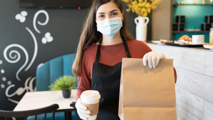 Woman wearing apron, mask, and rubber gloves, holding out paper bag and to-go coffee cup.