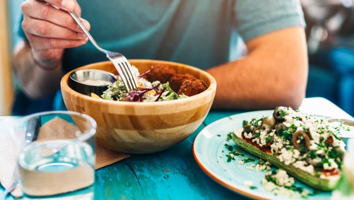 Shot of a young man eating fresh vegan salad with chickpeas balls and sesame sauce on a rustic wooden table.