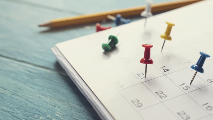 Calendar lying on a wooden surface with multi-colored push pins stuck to different dates and a pencil next to it.