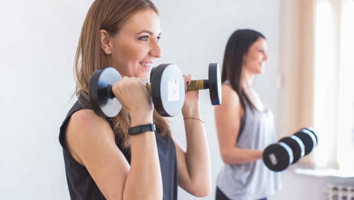 Two young women smiling and lifting dumbbells in a sunlit interior.