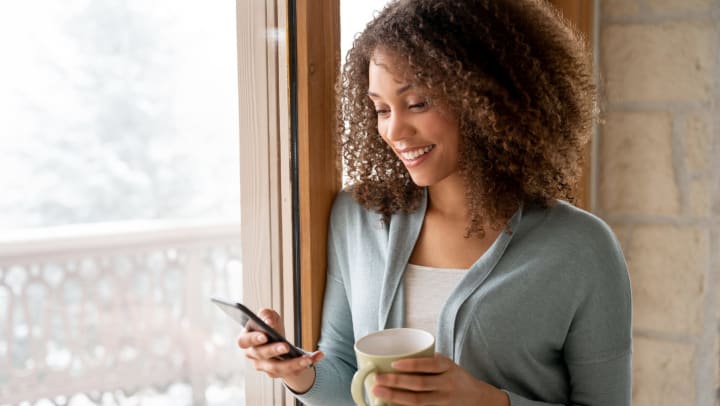 Woman leaning against a window frame while looking at her phone and holding a mug in her other hand. 