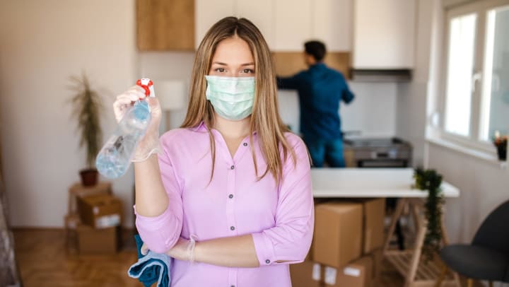 Woman wearing face mask and holding cleaning supplies with a man cleaning and boxes piled up in the background