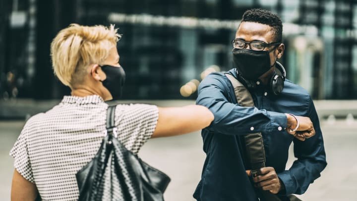 Two people outdoors, in masks, with buildings in the background, bumping elbows.