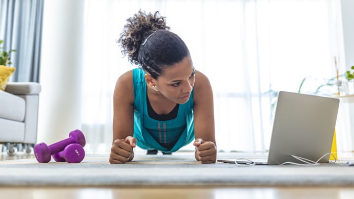 Low viewpoint of young woman in plank pose with dumbbells and laptop on the floor in a light-filled living room