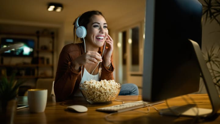 Young woman with headphones laughing while eating popcorn and watching a computer screen