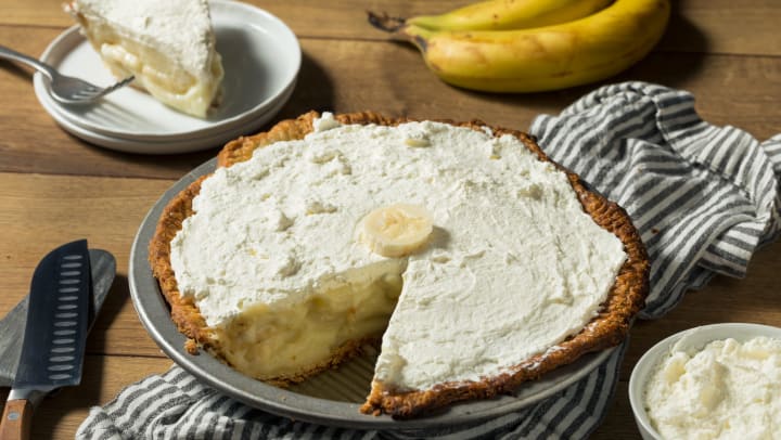 A homemade banana cream pie sits on a table, with one slice sitting on a plate behind it.