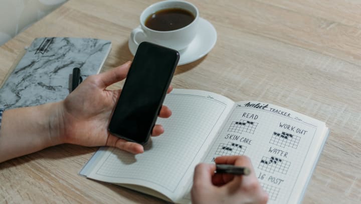 Person holding their phone and a pen with a habit tracker journal open on a table with an out of focus cup of coffee on it.