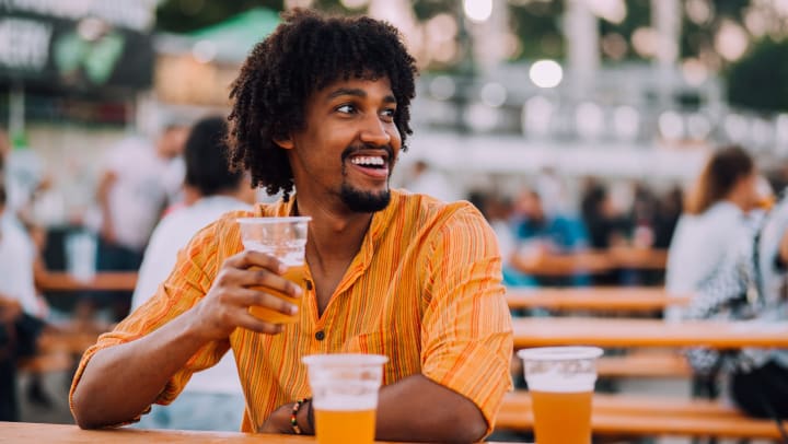 A smiling man sitting at a table on an outdoor patio while enjoying a beer.