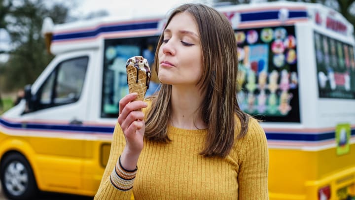 A woman standing in front of an icecream truck eating icecream in a waffle cone. 