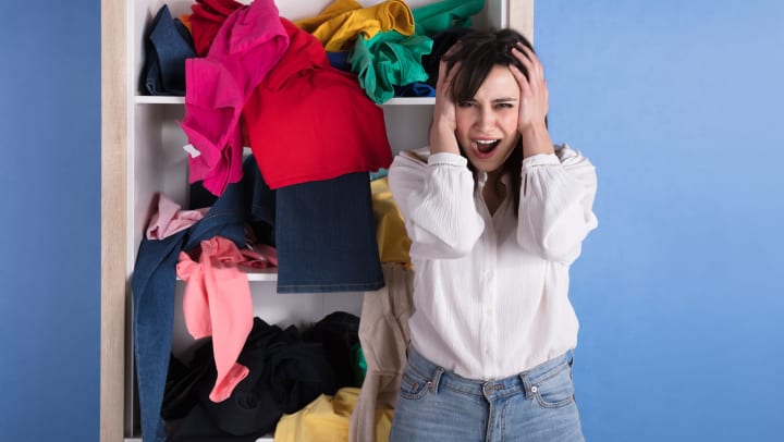 A stressed-looking young woman clutches her head with both hands as she stands in front of a messy closet.