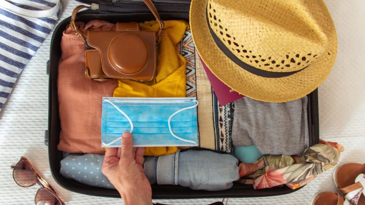 Hand putting a face mask in a suitcase. There are other accessories in the bag, including a hat, a camera, and a bikini.