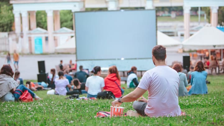 Group of people sitting on a lawn facing a large projector screen with a man in focus grabbing popcorn out of a bowl.