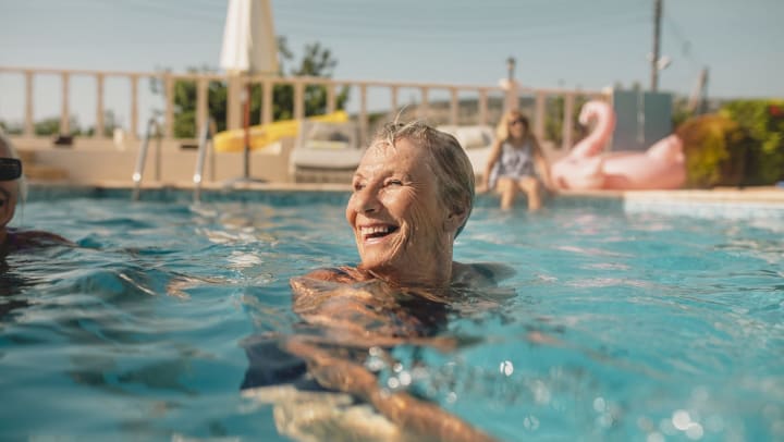 Elderly woman smiling in a pool with a person and pink flamingo behind her.