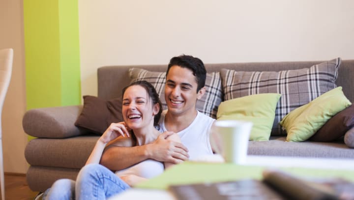 Young couple watching TV and smiling while sitting on the floor and leaning against a grey couch