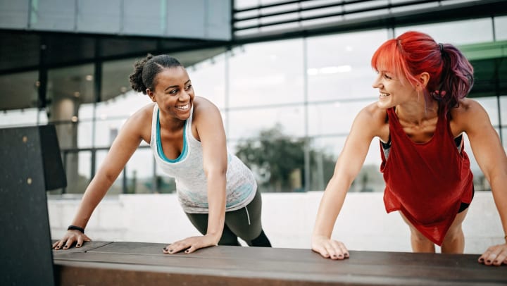 Two smiling women wearing athletic gear holding plank pose outside an office building