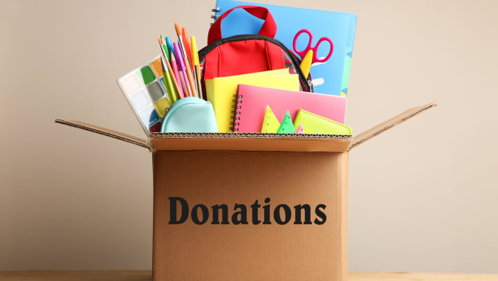 A bunch of school supplies sticking out of a brown cardboard box that says “Donations” on it. 