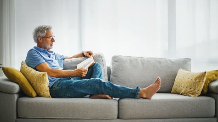 A relaxed man reading a book on the sofa in a well-lit room.