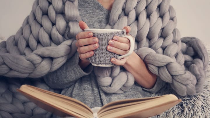 A woman with an oversized chunky knit blanket wrapped around her shoulders, holds a cup of coffee with a book open in her lap. 