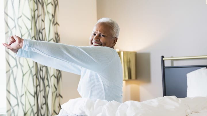 Smiling senior woman sitting up in bed and stretching in a light-filled bedroom